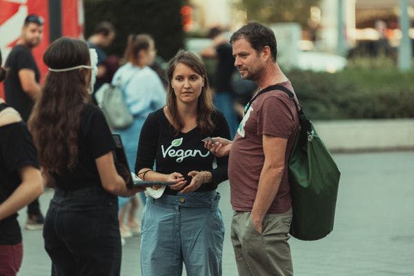 A képen két ember látható egy forgalmas utcán. Egy fiatal nő, aki egy fekete pólót visel, melyen a "Vegan" szó olvasható, beszélget egy középkorú férfival, aki egy barna pólót és egy zöld hátizsákot visel. A háttérben további emberek és városi környezet látható.